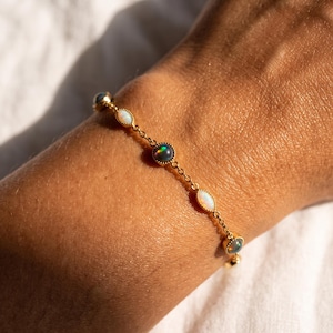 Könnte beinhalten: Ein goldenes Kettenarmband mit abwechselnd runden und marquiseförmigen Opalen. Die runden Opale zeigen dunkle Töne mit irisierenden Blitzen, während die marquiseförmigen Opale weiß mit subtilem Farbspiel sind, alle in goldenen Fassungen.