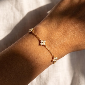 May include: A gold chain bracelet featuring three evenly spaced flower-shaped charms, each set with four small, iridescent white opal stones arranged in a clover pattern, connected by delicate gold links that wrap around the wrist.