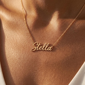Può includere: Una collana in metallo color oro caratterizzata da una catenina delicata e da un ciondolo centrale a forma di targhetta con il nome Stella scritto in un carattere corsivo fluido. Il ciondolo è appeso a una sottile catena a maglie lucide.