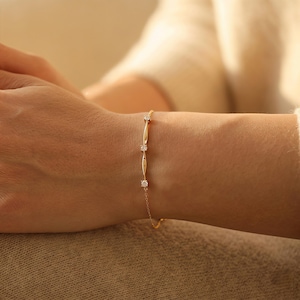 Può includere: Un delicato bracciale a catena in oro con tre piccole pietre rotonde e trasparenti incastonate nel metallo. Il design include sottili barre dorate allungate posizionate tra ogni pietra, creando un'estetica raffinata sul polso.