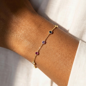 May include: A delicate gold chain bracelet featuring a series of small, round, bezel-set gemstones in various colours, including deep blue, light purple, and vibrant pink, spaced evenly along the thin, polished yellow gold metal link chain.