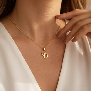 May include: A gold-colored necklace with a pendant featuring the intertwined letters "C" and "D". The necklace is suspended from a delicate chain. The pendant and chain appear to be made of a shiny metal.