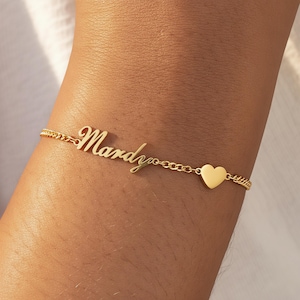 Puede incluir: Una pulsera dorada con el nombre "Mardy" en escritura cursiva. La pulsera presenta un pequeño dije de corazón y una delicada cadena. La pulsera se lleva en la muñeca.