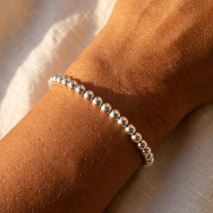 May include: A silver beaded bracelet. The bracelet is composed of small, round, shiny beads. The bracelet is worn on a wrist, with the skin tone appearing warm in the natural light. The background is a soft, off-white fabric.