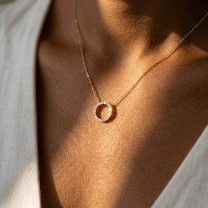 May include: A delicate gold necklace featuring a pendant in the shape of a circle, encrusted with small, sparkling gemstones. The necklace is worn against a skin tone, with sunlight casting shadows.