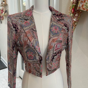 Może przedstawiać: Krótka marynarka z kolorowym wzorem paisley w odcieniach różu, błękitu i brązu. Kurtka ma długie rękawy i jest ozdobiona cekinami. Marynarka jest prezentowana na manekinie.