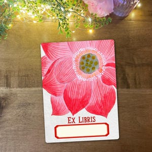 May include: A rectangular bookplate featuring a pink lotus flower watercolour illustration. The red text reads "EX LIBRIS", with a blank space for customisation. The background is a wooden surface.