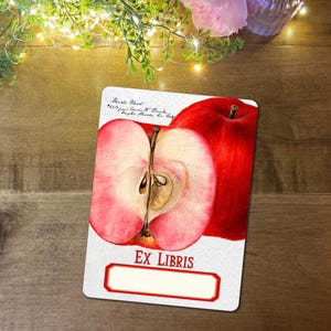 May include: A rectangular Ex Libris card with a watercolour illustration of a red apple, one half sliced to show the core. The card has a red border and the words "EX LIBRIS" at the bottom. The background is a wooden surface.