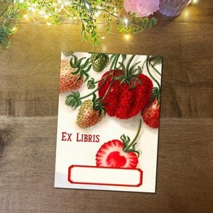 May include: A white Ex Libris bookplate with a watercolour illustration of strawberries. The design includes ripe red strawberries and unripe green ones, with the text "EX LIBRIS" in red. A blank label is at the bottom.