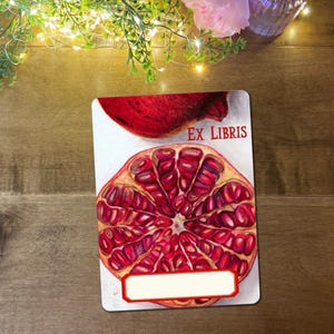 May include: A rectangular bookplate with a detailed illustration of a pomegranate, and the words "EX LIBRIS" in red. It has a space for personalization. The bookplate is set against a wooden background.