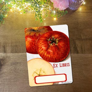 May include: A rectangular bookplate with a watercolour illustration of red apples. The bookplate has the words "EX LIBRIS" in red, with a blank space for a name. The bookplate is on a wooden surface.