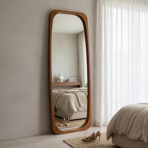 May include: A full-length mirror with a wooden frame and rounded edges. The mirror is positioned against a light grey wall in a bedroom. The frame has a natural wood finish. The mirror reflects a bed, a small table, and a window with white curtains.