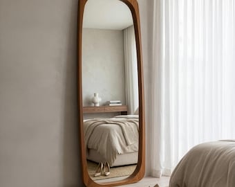 May include: A tall, rectangular wooden-framed mirror with rounded corners. The mirror is leaning against a light gray wall in a bedroom setting. The frame has a natural wood finish. The mirror reflects a bed, a small table, and a window with white curtains.