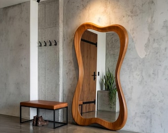 May include: A large, wooden-framed mirror with an organic, hourglass shape leans against a concrete wall. A brown leather bench with a black metal frame and a pair of brown boots sit nearby. The room has a modern, minimalist design.