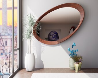 May include: Oval wooden-framed mirror on a white wall. The mirror reflects a room with a painting and a window. A tall white vase with a plant and small vases with flowers are in front of the wall.