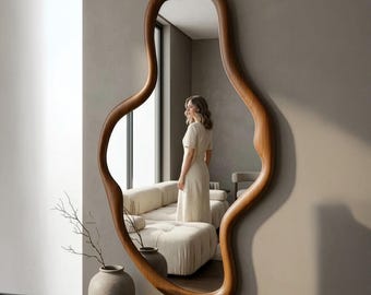 May include: A large, organically shaped wooden mirror with a warm brown finish hangs on a neutral-toned wall. The mirror's unique frame curves and flows, reflecting a room with a sofa, chair, and a person. A vase with branches sits below.