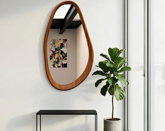 May include: A large, organic-shaped wooden mirror hangs on a white wall. The mirror's frame is a warm brown color. Below the mirror is a black bench with a minimalist design. A potted plant with large green leaves stands to the right.