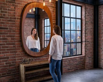 May include: A woman stands before a large, oval wooden mirror in a room with exposed brick walls and a city view. The mirror is mounted on a wooden console table. Decorative light bulbs hang from the ceiling.