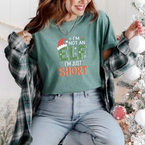 Pode incluir: Uma camiseta verde com o texto "I'M NOT AN ELF I'M JUST SHORT" em branco, verde e laranja. O design inclui um gorro de Papai Noel e flocos de neve. A pessoa está vestindo uma camisa xadrez verde e preta e jeans azuis.