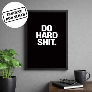 May include: Black framed wall art with the text "DO HARD SHIT" in bold white letters. A white circle with the words "INSTANT DOWNLOAD" is in the upper left corner. A black coffee mug, glasses, and a notebook are on a wooden surface.
