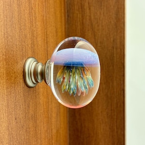 Puede incluir: Un pomo de armario de cristal transparente con un diseño abstracto y colorido en su interior. El pomo tiene una base de color plateado y está montado en una puerta de armario de madera. El diseño interior del pomo presenta tonos azules, amarillos y naranjas.