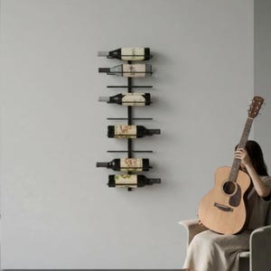 May include: A black metal wine rack mounted on a light gray wall, holding six bottles of wine. The bottles are dark with white and beige labels. A person is holding a wooden acoustic guitar.