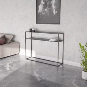 May include: A dark grey metal console table with two shelves. The table has a rectangular frame and is against a light grey wall. Decorative items and a stack of magazines are on the shelves. A framed abstract artwork hangs above the table.