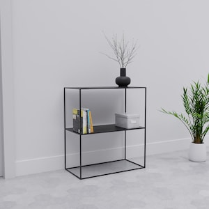 Op de afbeelding: Zwart metalen rek met twee planken. De bovenste plank bevat een zwarte vaas met takken en de onderste plank boeken en een grijze doos. Een plant in een pot staat rechts.