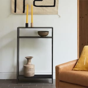 Puede incluir: Estantería de metal negro con dos estantes, con un cuenco de madera, un jarrón decorativo y dos velas amarillas. Un tapiz de pared tejido de color beige y un sillón de cuero están en el fondo.