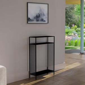 Op de afbeelding: Een zwart metalen rek met twee planken en een gaas zijpaneel. Het rek staat tegen een lichtgekleurde muur, met een ingelijst abstract kunstwerk erboven. De kamer heeft een houten vloer en uitzicht op een tuin.