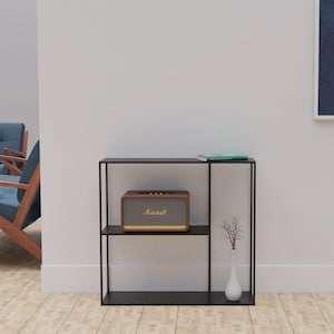 May include: A black metal shelf unit with three levels. A brown speaker with the name "Marshall" is on the middle shelf. A small white vase with a decorative branch is on the bottom shelf. A book rests on the top shelf.