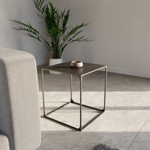 Pode incluir: Uma mesa lateral quadrada moderna com um tampo cinza escuro e uma estrutura de metal preta. A mesa tem 50 cm de lado e tem um design minimalista. Uma pequena tigela clara está em cima. Uma planta em vaso está ao fundo.