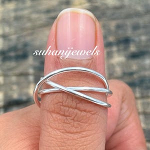 Puede incluir: Un anillo de plata con un diseño entrecruzado único. El anillo presenta múltiples bandas finas que se cruzan, creando un aspecto abierto y moderno. El anillo se muestra en un dedo, con el nombre de la marca "suhanijewels" visible.