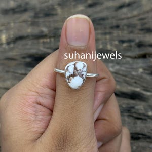 May include: A silver ring with a teardrop-shaped white turquoise stone with brown veining. The ring is set on a silver band. The text "suhanijewels" is visible on the image.