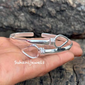 May include: A silver cuff bracelet with a unique design. The bracelet features two open teardrop shapes connected by a wrapped wire design. The brand name "Suhanijewels" is printed on the bracelet.
