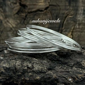 Op de afbeelding: Een stapel zilveren armbanden, gepresenteerd op een getextureerd, donkerbruin oppervlak. De armbanden zijn dun en glanzend en reflecteren licht. De tekst "suhanijewels" is rechtsboven zichtbaar.