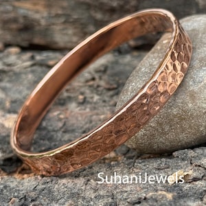 May include: A copper-colored bangle bracelet with a hammered texture. The bracelet is round and has a smooth interior. The surface of the bracelet has a textured, dimpled pattern. The text "SuhaniJewels" is visible on the surface.