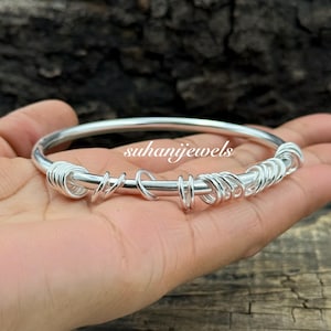 May include: A silver bangle bracelet with a minimalist design. The bracelet has a smooth, rounded band with decorative coiled and looped wire accents. The text "suhanijewels" is visible on the bracelet. The jewelry is held in a hand.
