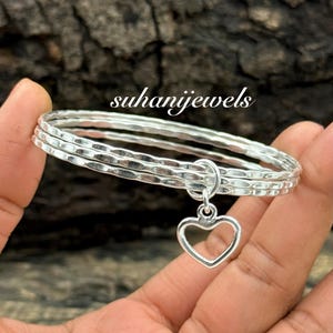 Op de afbeelding: Een zilveren armbandenset met drie gehamerde banden en een hangende hartvormige bedel. De armband wordt in een hand gehouden tegen een vage houten achtergrond. De tekst "suhanijewels" is zichtbaar.