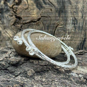 Puede incluir: Dos pulseras de plata con un acabado texturizado y detalles florales descansan sobre una piedra lisa y redondeada. Las pulseras se exhiben sobre un fondo de madera oscura y rústica. El texto "Suhanijewels" es visible.