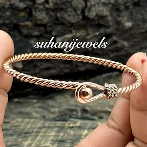 May include: A rose gold twisted wire bangle bracelet with a hook and eye closure. The bracelet is held in a person's hand. The text "suhanijewels" is in the image.