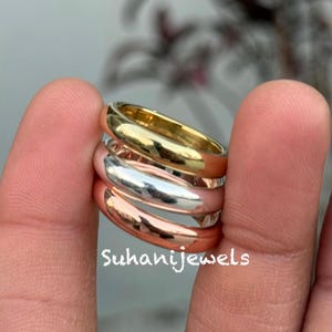 Könnte beinhalten: Ein Stapel von vier Ringen in Gold-, Silber- und Roségoldtönen. Die Ringe sind glatt und glänzend, mit einem klassischen Banddesign. Die Ringe werden zwischen zwei Fingern gehalten. Der Text "Suhanijewels" ist sichtbar.