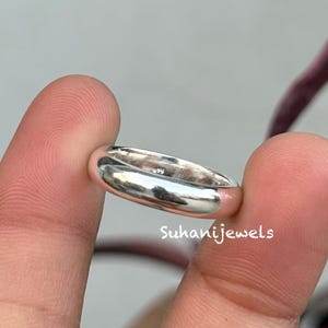 Könnte beinhalten: Ein polierter Silberring mit einem glatten, abgerundeten Band. Der Ring wird zwischen zwei Fingern gehalten, mit dem Markennamen "Suhanijewels" in weißem Text. Die Oberfläche des Rings reflektiert das Licht und hebt sein glänzendes Finish hervor.