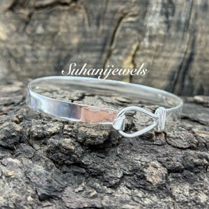 May include: A silver bangle bracelet with a hook clasp. The bracelet is made of polished metal and has a simple, elegant design. The text "Suhanijewels" is in the image.
