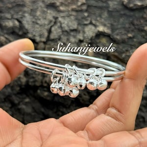 Può includere: Braccialetti d'argento con piccoli ciondoli a forma di campana pendenti. I braccialetti sono tenuti in una mano, con il testo "Suhanijewels" sullo sfondo. Le campane sono rotonde e lucenti, aggiungendo un tocco delicato ai gioielli.