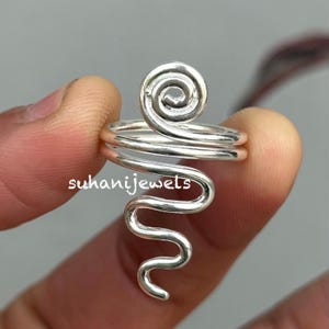 Puede incluir: Un anillo plateado con un diseño en espiral en la parte superior y una forma ondulada y alargada. El anillo está sujeto por una persona. El texto "suhanijewels" es visible en el anillo.