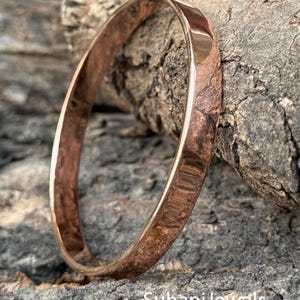 Peut inclure: Un bracelet jonc de couleur cuivrée avec une surface lisse et polie. Le bracelet est circulaire et semble être en métal. Le fond est une surface texturée, grise et brune. Le texte "SuhaniJewels" est visible en bas.
