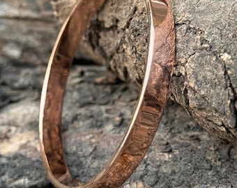 May include: A copper-toned bangle bracelet with a smooth, polished finish. The circular bracelet is made of metal. The background is a textured, grey and brown surface. The text "SuhaniJewels" is visible at the bottom.