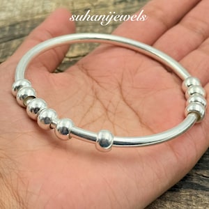 May include: A silver bangle bracelet featuring a series of spherical beads. The bracelet is held in a hand, with the brand name "suhanijewels" visible. The design is simple and elegant.
