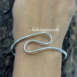Könnte beinhalten: Ein silberfarbenes Armreif-Armband mit einem fließenden, offenen Design. Das Armband umschließt das Handgelenk mit einer glatten, polierten Oberfläche. Der Text "Suhanijewels" ist sichtbar.